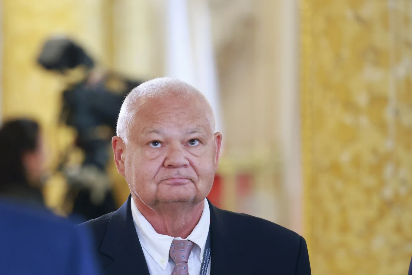 RPP może dokonać obniżki stóp procentowych już na październikowym posiedzeniu. Na zdj. przewodniczący RPP i prezes NBP Adam Glapiński Mężczyzna w garniturze i krawacie na eleganckim tle o złocistych odcieniach, w tle rozmyta postać i kamera, sugerujące oficjalne wydarzenie lub konferencję.