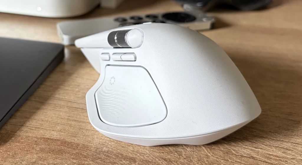 Logitech MX Master 4 wprowadza zmiany obejmujące dizajn oraz użyte materiały. Biała mysz komputerowa z przyciskami bocznymi ustawiona na drewnianym blacie obok smartfona i innych akcesoriów elektronicznych.