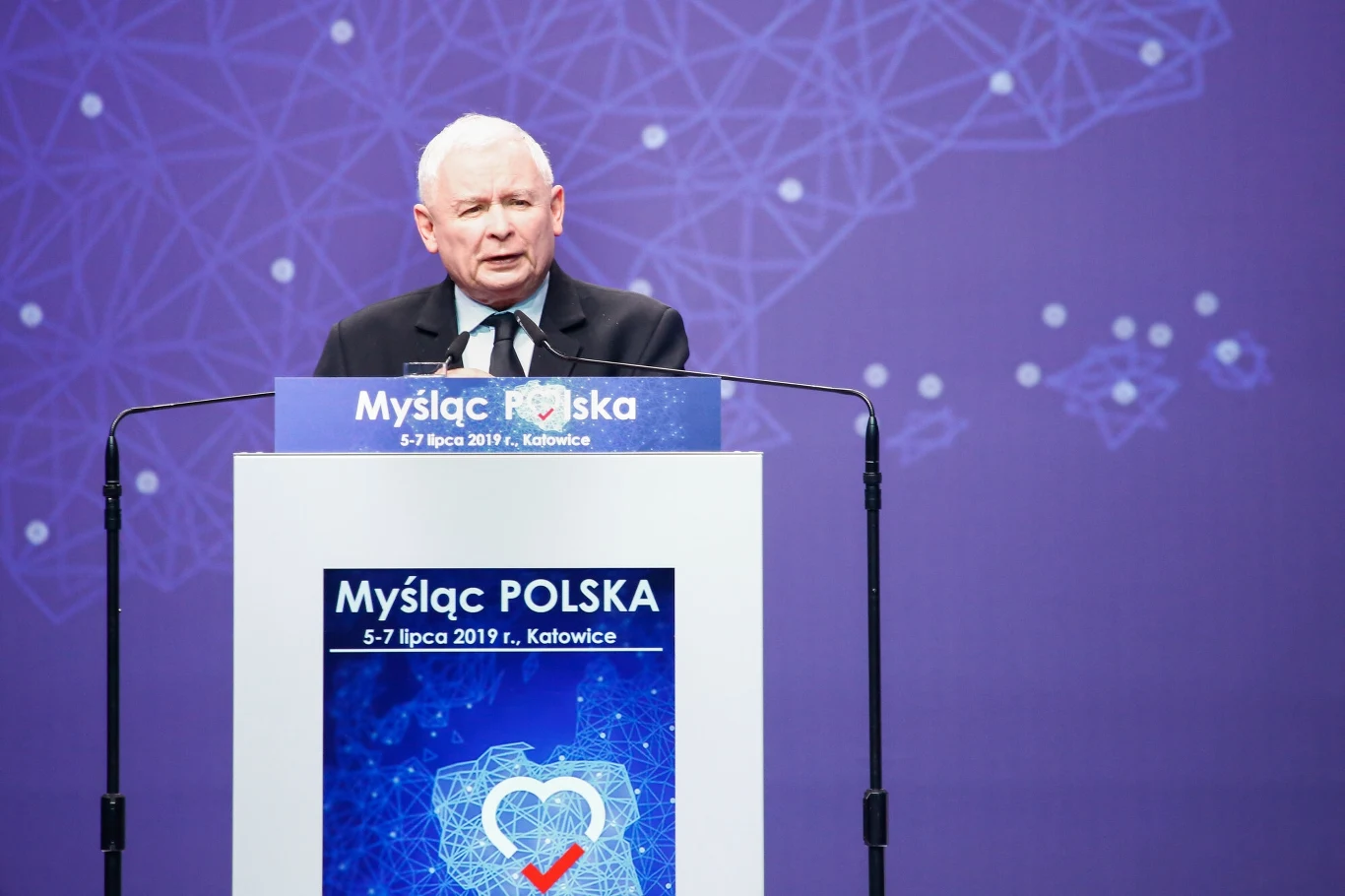 Jarosław Kaczyński podczas konwencji programowej w Katowicach w 2019 roku Starszy mężczyzna w garniturze przemawia zza mównicy podczas konferencji Myśląc Polska, z tłem w odcieniach niebieskiego przedstawiającym graficzną mapę sieci i serce z zaznaczeniem.