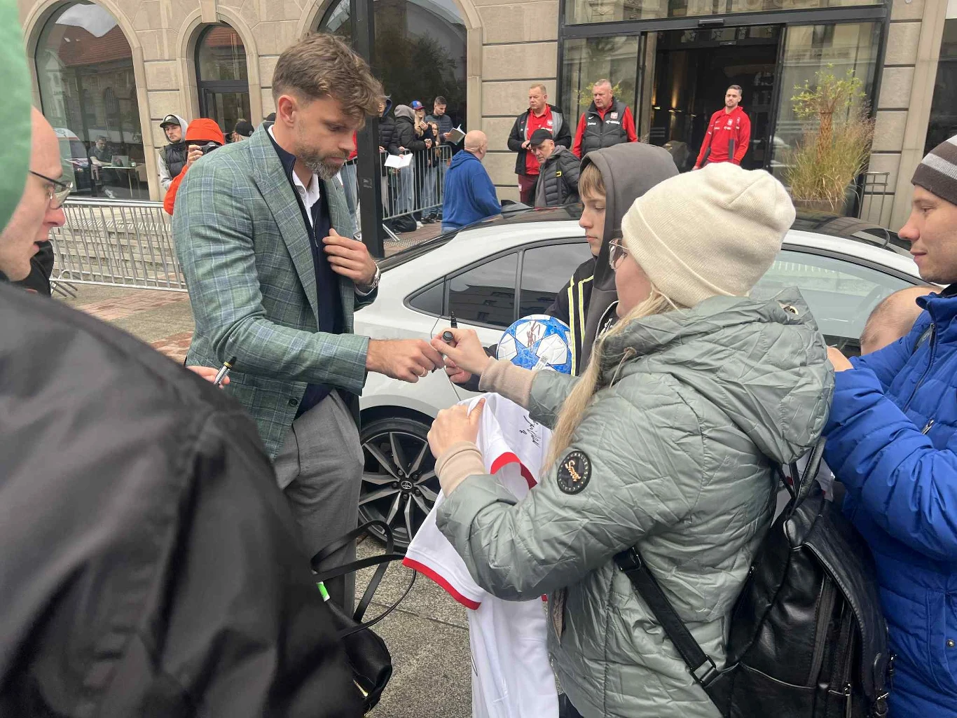 Mężczyzna w eleganckim ubraniu rozdaje autografy grupie fanów, jedna z osób trzyma piłkę i koszulkę sportową. W tle zaparkowany samochód oraz kilka osób obserwujących sytuację.