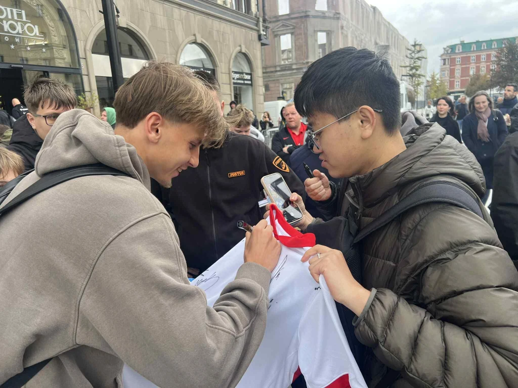 Jan Ziółkowski Młody mężczyzna podpisuje koszulkę innemu młodemu mężczyźnie, wokół stoją ludzie, w tle widoczne miasto i budynki.
