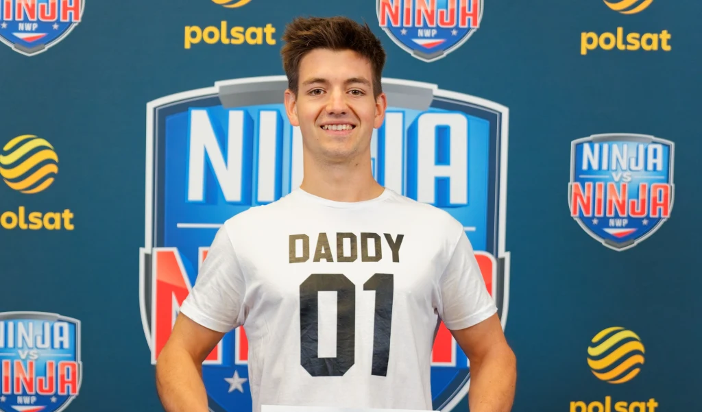 Jan Tatarowicz w ostatnim odcinku eliminacyjnym „Ninja vs Ninja” Młody mężczyzna w białej koszulce z napisem 'DADDY 01' stoi na tle ścianki z logotypami Polsat oraz Ninja vs Ninja, uśmiecha się do kamery.