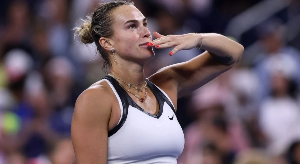 Tenisistka w sportowym stroju wysyła pocałunek w stronę widowni, w tle rozmazana publiczność podczas zawodów tenisowych.