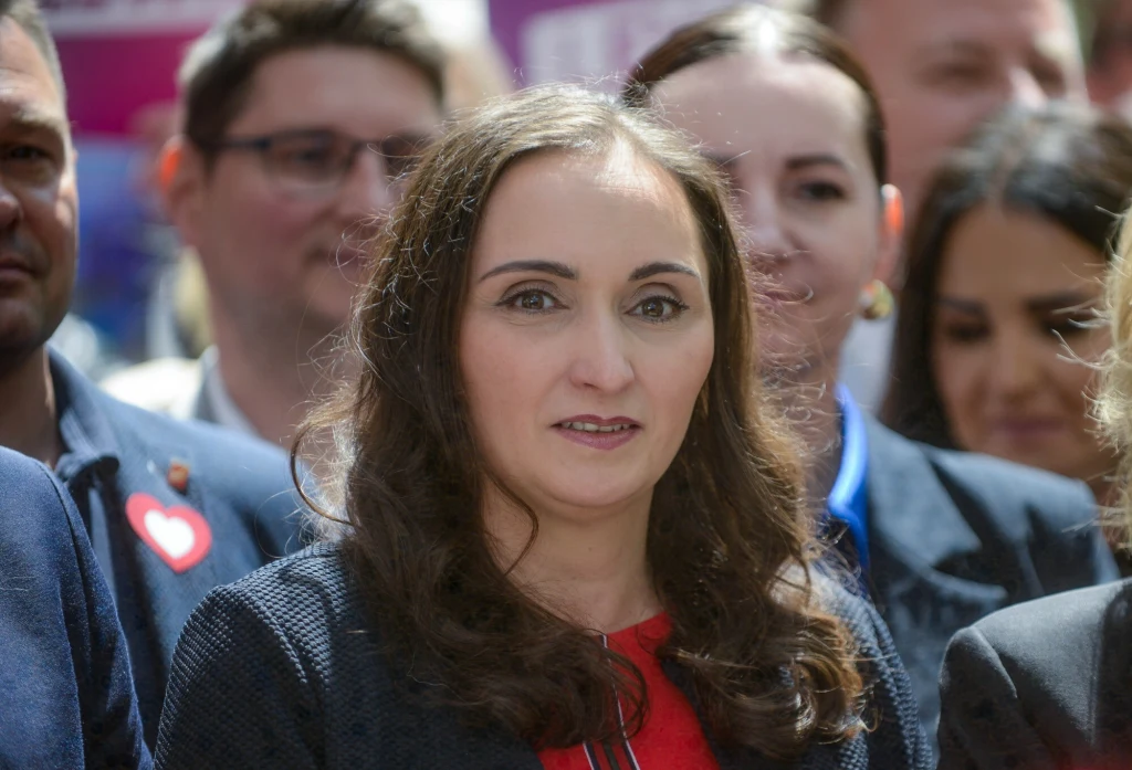 Ewa Szymanowska Kobieta z długimi ciemnymi włosami na pierwszym planie ubrana w czerwony strój oraz ciemną marynarkę, wokół niej grupa ludzi, scena prawdopodobnie w plenerze, atmosfera oficjalna lub społeczna.