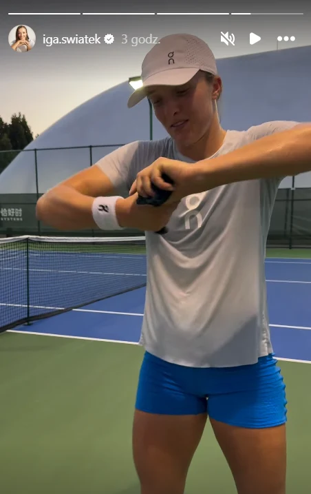 Kobieta w sportowym stroju na korcie tenisowym, ubrana w białą koszulkę i niebieskie spodenki, trzyma w rękach przyrząd przypominający owijkę do rakiety lub inny akcesorium tenisowy, w tle widoczna siatka i linie kortu.