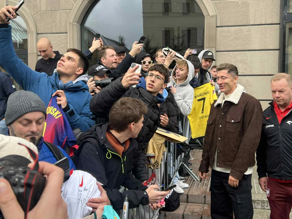 Robert Lewandowski Grupa kibiców otacza znaną osobę stojącą przy barierkach, robiąc z nią selfie oraz trzymając piłkarskie koszulki i gadżety do podpisu; osoby są ubrane w kurtki, na jednym z gadżetów widoczny napis 'Lewandowski'.