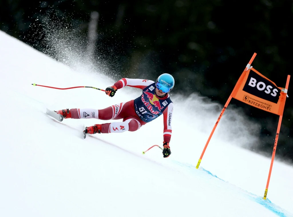 Julian Schuetter Narciarz w pełnym stroju sportowym z niebieskim kaskiem dynamicznie pokonuje zakręt na trasie slalomu giganta, obok tyczki oznaczonej flagą BOSS, na tle białego śniegu i rozmytego, ciemnego lasu w tle.