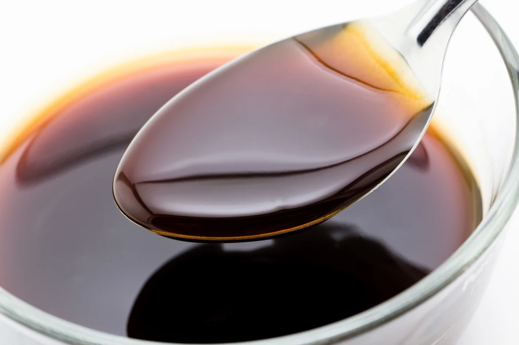 Srebrna łyżka z ciemnobrązowym płynem nad przezroczystą miseczką wypełnioną tym samym płynem, całość utrzymana w jasnej kolorystyce tła