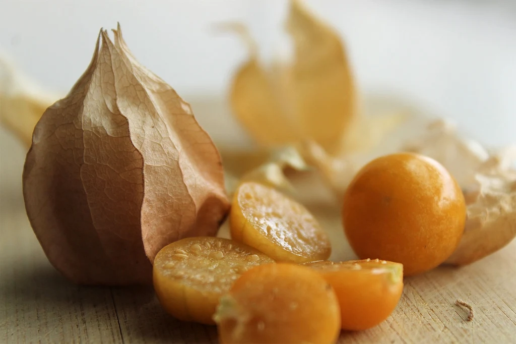 Pomarańczowe owoce physalis na drewnianej powierzchni, część z nich jest w całości, część przekrojona, a w tle widoczne fragmenty uschniętej łuski.