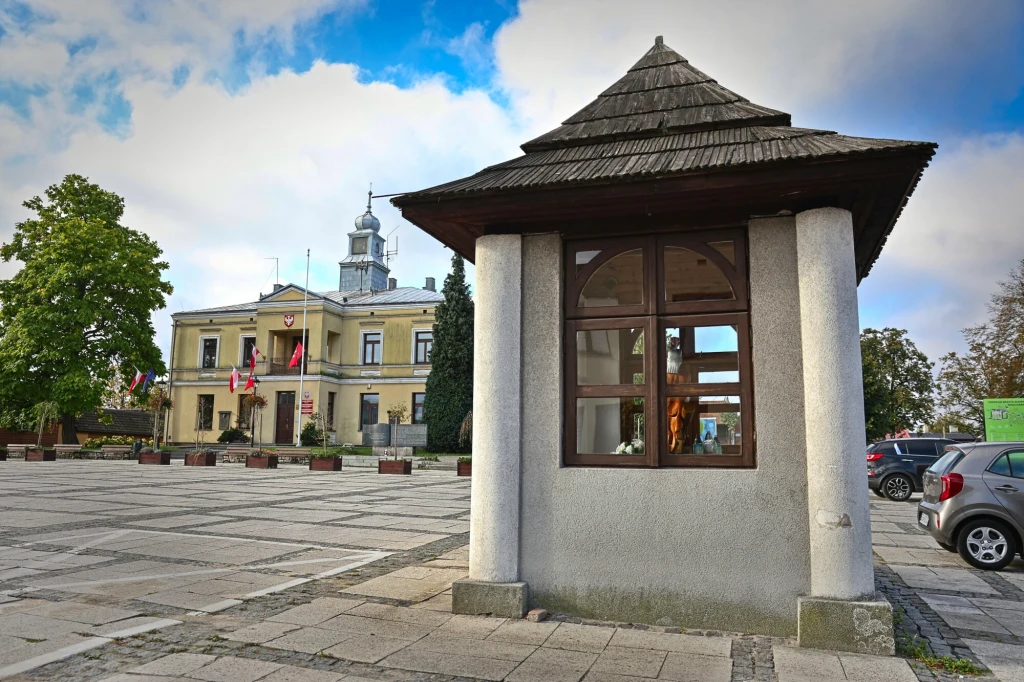 Kamienny kiosk z drewnianym dachem na pierwszym planie stojący na wyłożonym płytkami rynku, w tle zabytkowy budynek ratusza z wieżą zegarową i flagami, po prawej zaparkowany samochód.
