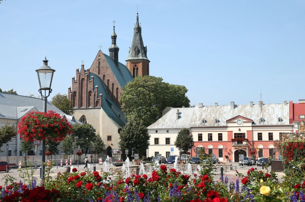 Historyczny rynek z kwietnikiem na pierwszym planie, w tle zabytkowy kościół z wysokimi wieżami i charakterystyczną, ceglano-gotycką architekturą, otoczony starymi budynkami i drzewami.