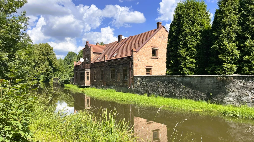 Ceglany budynek o spadzistym dachu położony nad kanałem wodnym, otoczony zielenią i wysokimi drzewami, w słoneczny dzień z niebem pokrytym białymi chmurami.