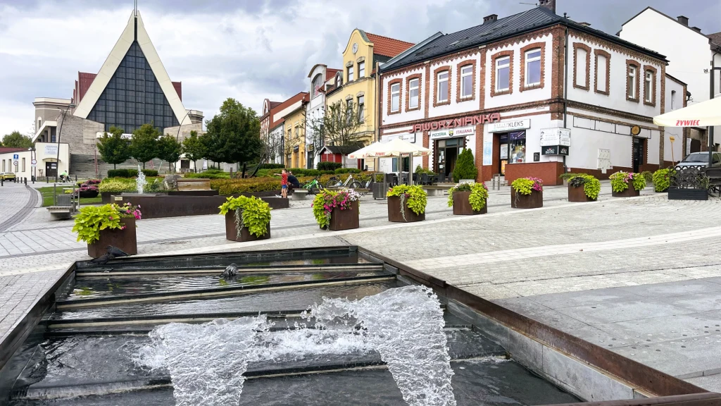 Plac miejski z fontanną na pierwszym planie, otoczony zadbaną zielenią w donicach, z nowoczesnym kościołem i kilkoma kamienicami z lokalami użytkowymi po bokach.