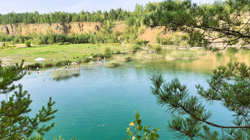 Lazurowa woda otoczona zielenią, stromy klif w tle, na brzegu ludzie odpoczywają oraz spacerują po drewnianych pomostach, pierwszoplanowe gałęzie sosny podkreślają naturalny charakter miejsca