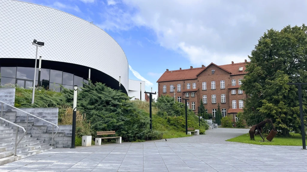 Nowoczesny, owalny budynek z białą fasadą sąsiaduje z klasycznym, czerwonym, ceglastym gmachem pośród zieleni, przy szerokim placu wyłożonym szarymi płytami, ze schodami i rzeźbą konia na trawie.