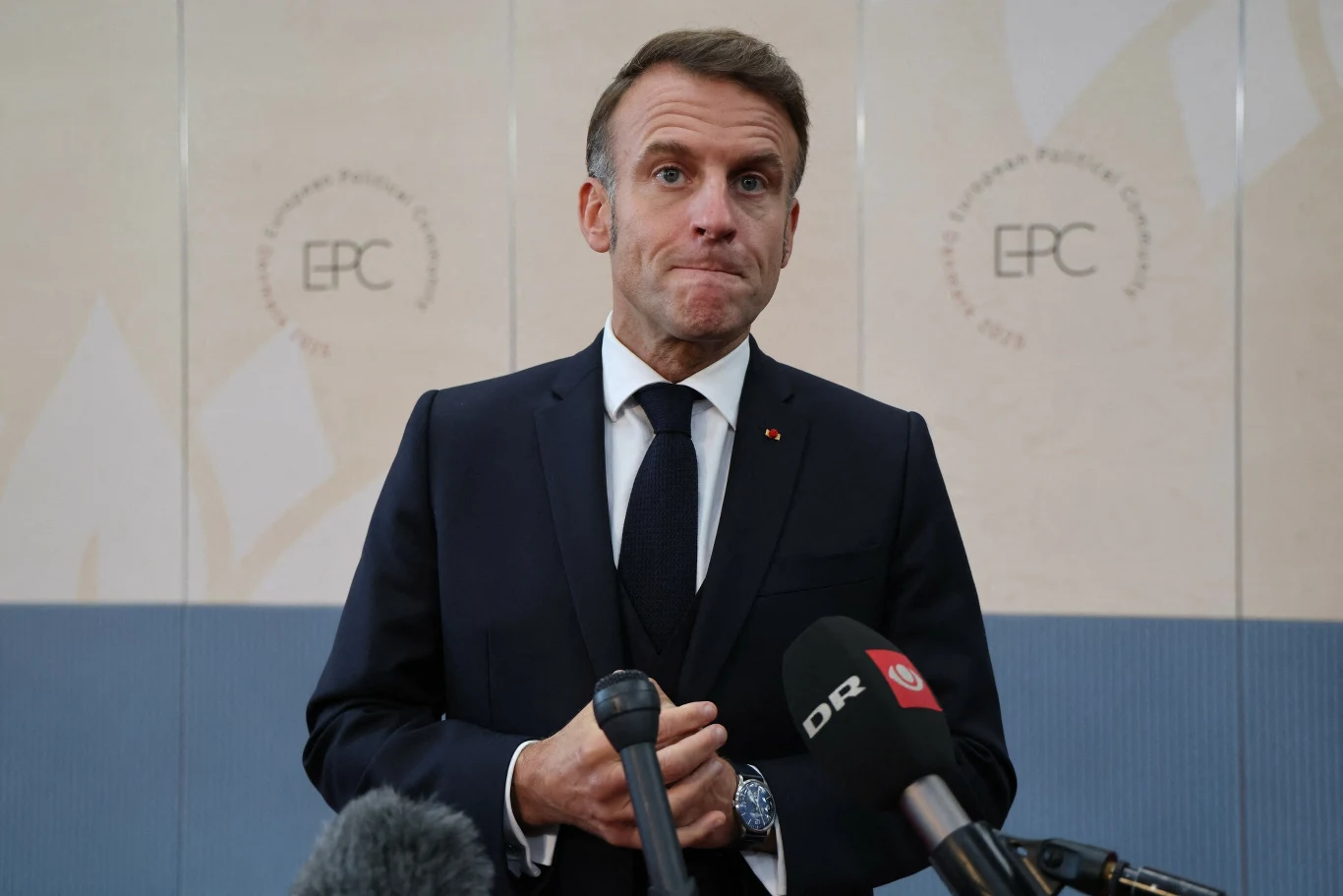 Prezydent Francji Emmanuel Macron Mężczyzna w garniturze i krawacie stoi przy mikrofonach podczas konferencji prasowej, w tle widoczne logo EPC oraz neutralna, jasna ściana.