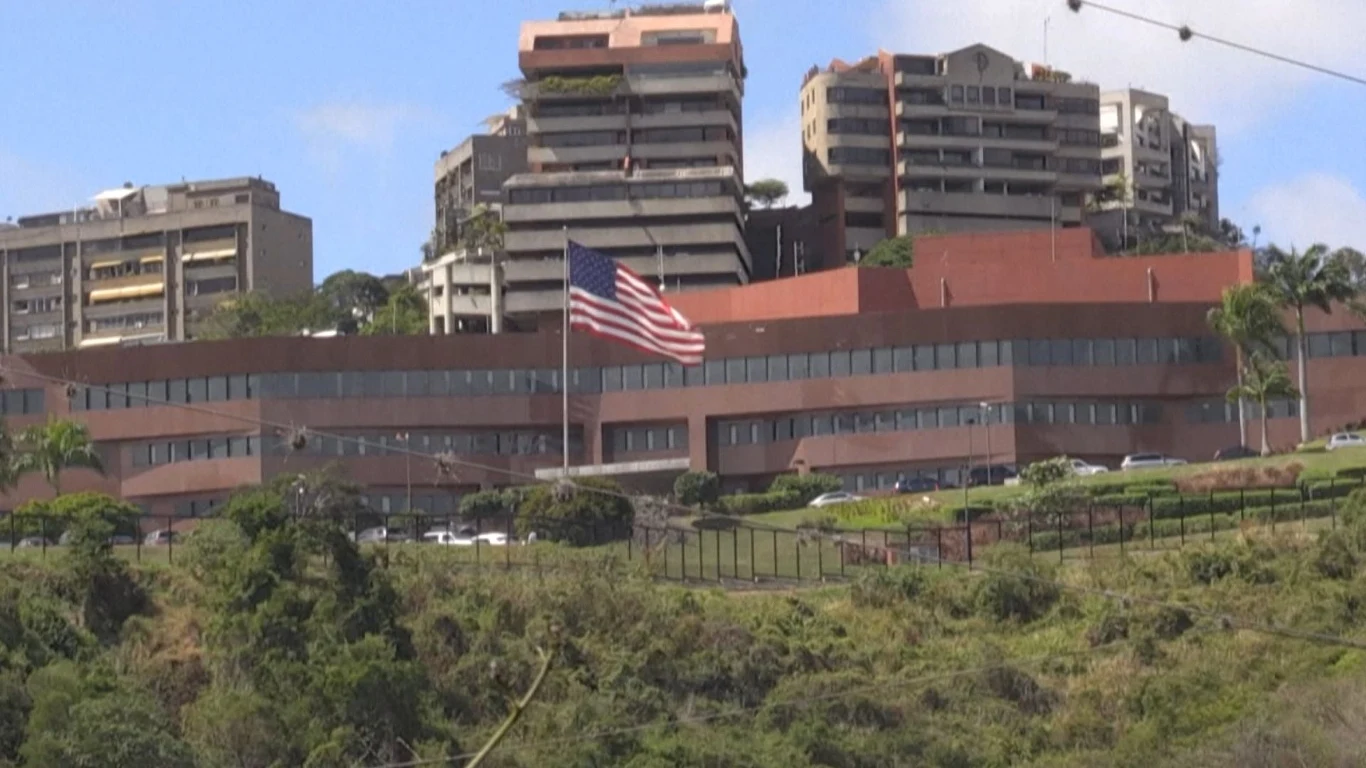 Ambasada USA w Caracas w Wenezueli Budynek o nowoczesnej architekturze z widoczną flagą Stanów Zjednoczonych na masztcie, otoczony zielenią i pobliskimi zabudowaniami, ustawiony na wzniesieniu, z drogą prowadzącą wzdłuż terenu.