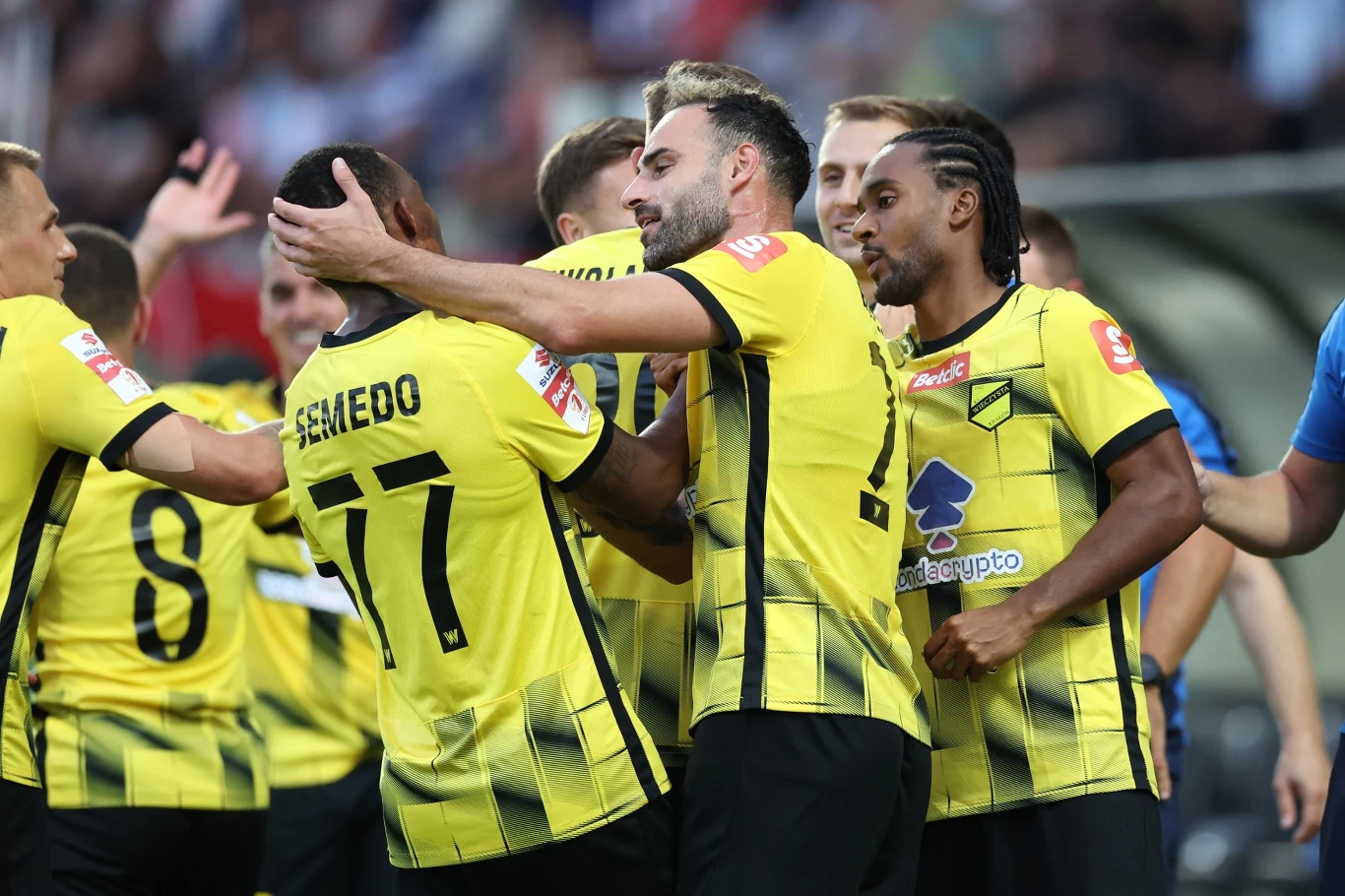 Grupa piłkarzy w żółto-czarnych strojach drużynowych świętuje zdobycie bramki na boisku, okazując emocje poprzez przytulanie się i radość, widoczne są nazwiska graczy na koszulkach, tłum w tle.