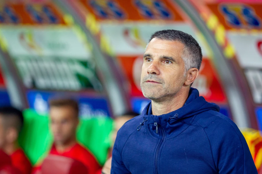 Gino Lettieri Mężczyzna w średnim wieku w granatowej bluzie z kapturem stoi w przestrzeni stadionowej, na tle kolorowych siedzeń i rozmytych postaci siedzących w ławce rezerwowych.