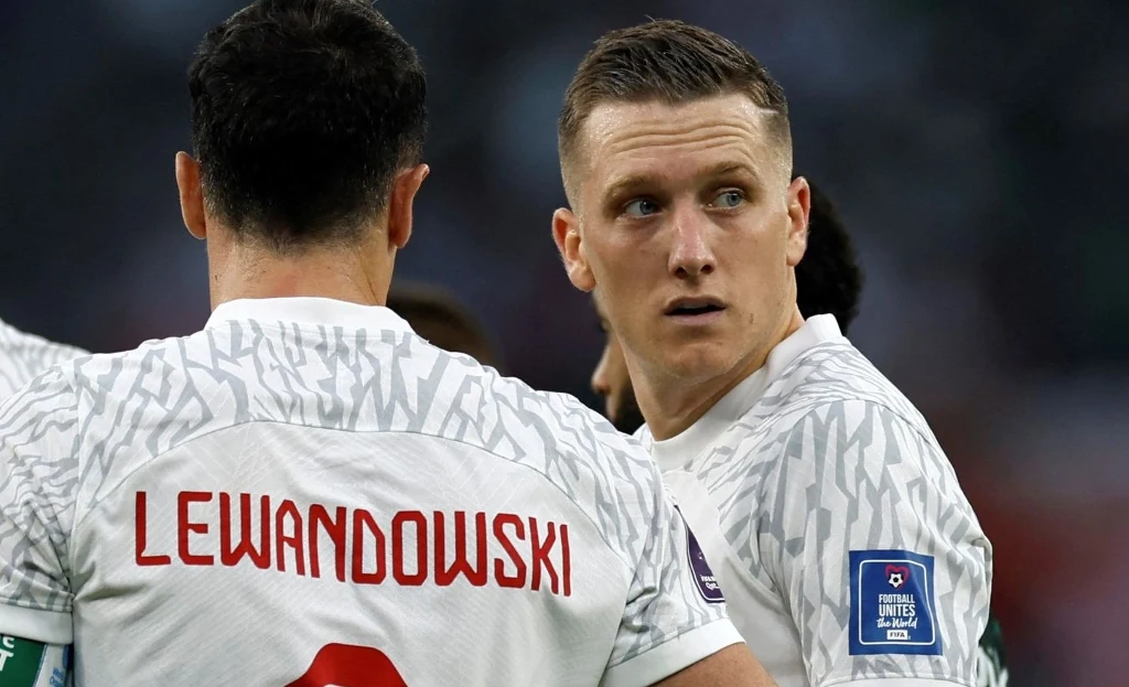 Piłkarze ubrani w biało-szare stroje sportowe podczas meczu, jeden z nich z widocznym nazwiskiem na plecach, w tle rozmyte postacie i stadionowe światło.