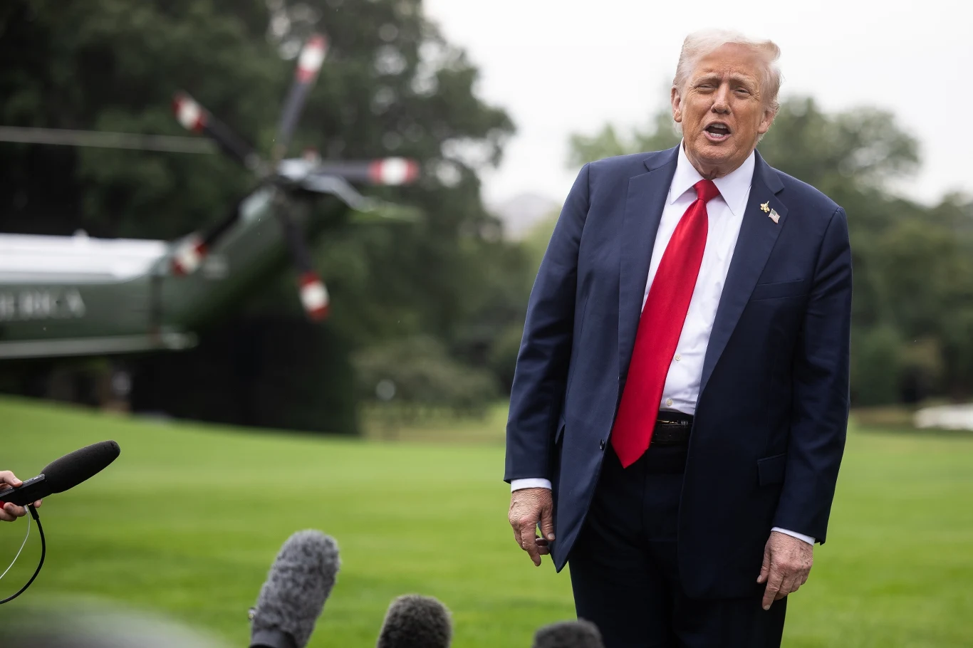 Donald Trump Mężczyzna w garniturze z czerwonym krawatem stoi na trawniku, za nim widoczny helikopter i mikrofony nagrywające wypowiedź.