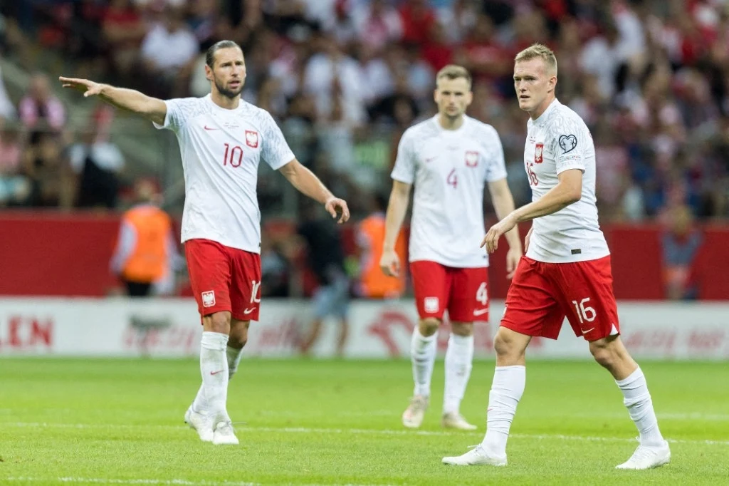 Grzegorz Krychowiak Polscy piłkarze w trakcie meczu, ubrani w białe koszulki z czerwonymi elementami i czerwone spodenki, na tle stadionu wypełnionego kibicami.