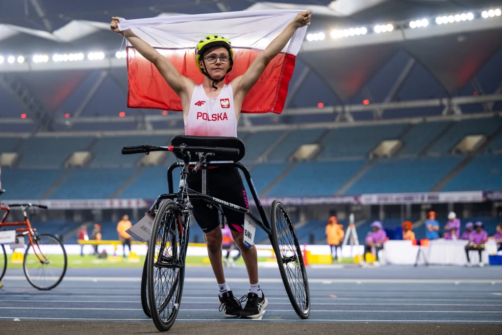 Sportowiec poruszający się na specjalnym rowerze trzyma polską flagę uniesioną nad głową na tle stadionu lekkoatletycznego. Na stroju zawodnika widnieje napis 'Polska' oraz godło narodowe.