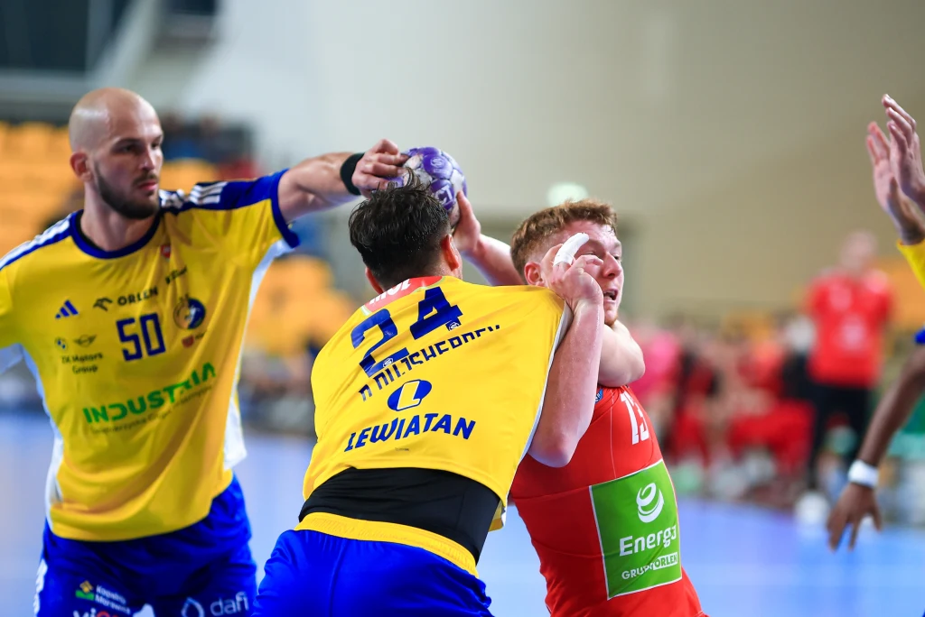 Zawodnik drużyny żółto-niebieskiej próbuje zatrzymać zawodnika drużyny czerwonej podczas meczu piłki ręcznej, trzymając go za twarz ręką, z boku widoczny kolejny zawodnik obserwujący sytuację, w tle ławka rezerwowych.