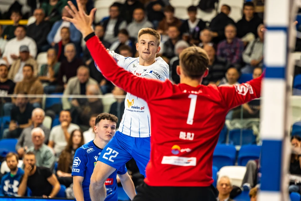 Zawodnik w białej koszulce wykonuje atak na bramkę podczas meczu piłki ręcznej, stojąc tuż przed bramkarzem w czerwonym stroju, a w tle widoczna jest publiczność na trybunach.