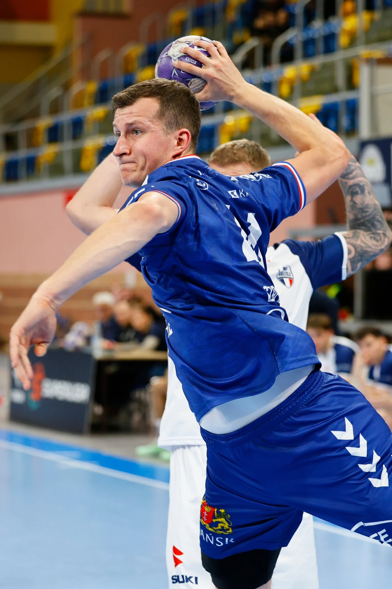 Mikołaj Czapliński, skrzydłowy PGE Wybrzeża Gdańsk Zawodnik w niebieskim stroju sportowym unosi się w powietrzu, trzymając piłkę ręczną w jednej dłoni, podczas akcji meczu rozgrywanego w hali sportowej, w tle widoczni inni gracze i trybuny.