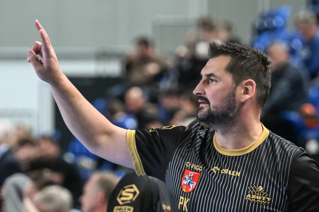 Patryk Kuchczyński, trener Azotów Puławy Mężczyzna w sportowej koszulce z herbem drużyny i napisem 'VIGOR ORLEN', wskazujący palcem w górę, tło rozmyte z siedzącymi ludźmi na widowni.