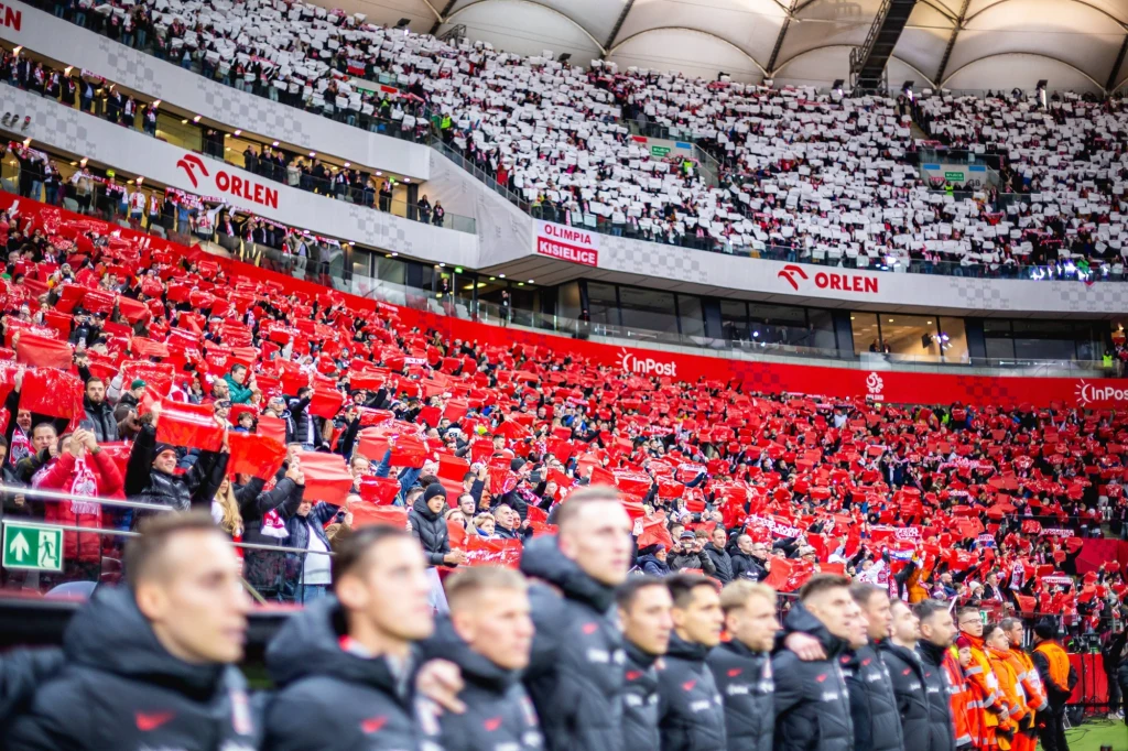 Tłum kibiców ubranych w czerwone i białe barwy narodowe na stadionie, tworzących flagę Polski; na pierwszym planie widoczna jedenastka piłkarska stojąca ramię w ramię