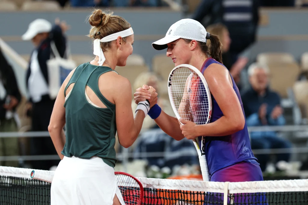 Dwie tenisistki ściskają sobie dłonie przy siatce po zakończonym meczu na korcie, obie ubrane w sportowe stroje, w tle widoczni rozmyci kibice na trybunach.