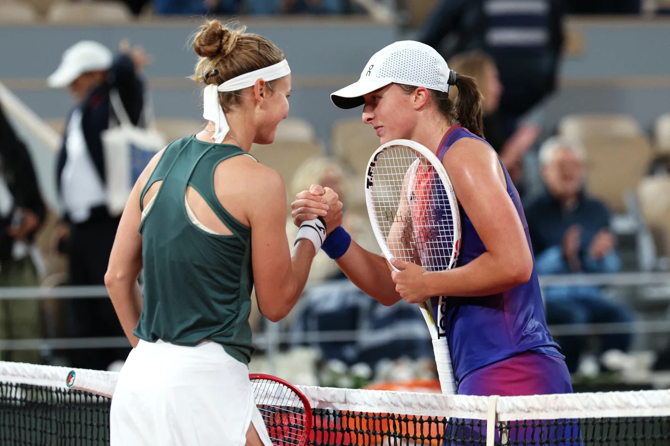 Dwie tenisistki ściskają sobie dłonie przy siatce po zakończonym meczu na korcie, obie ubrane w sportowe stroje, w tle widoczni rozmyci kibice na trybunach.