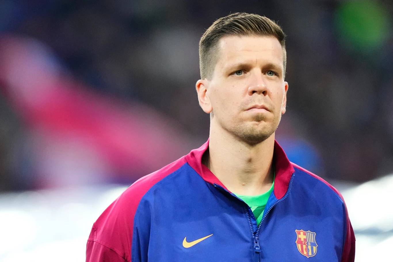 Mężczyzna w sportowej kurtce w barwach FC Barcelony stoi na stadionie, skupiony i poważny, z rozmytym tłem trybun.