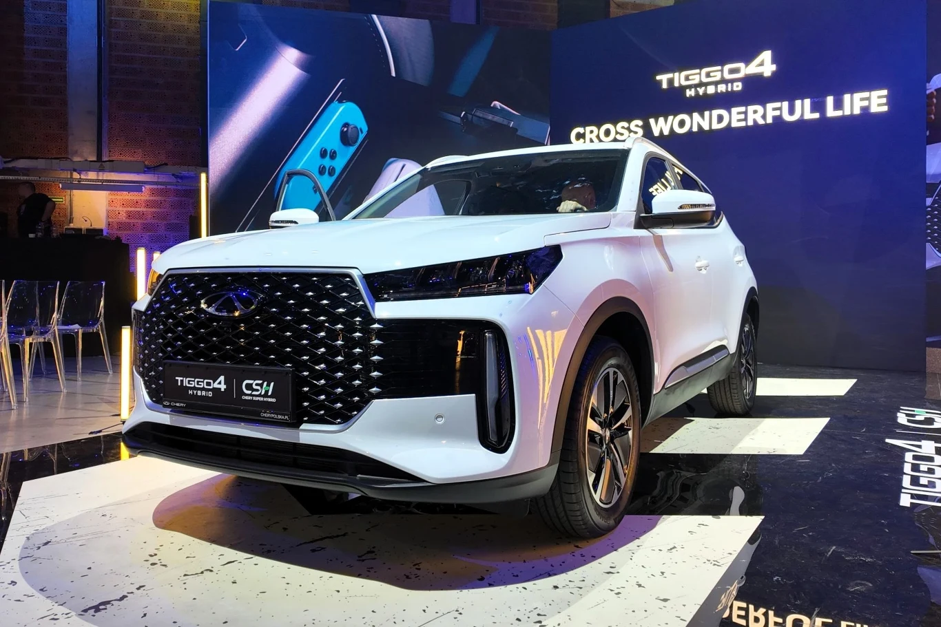 Chery Tiggo 4 HEV dołącza do polskiej oferty. Wystarczy 95 900 zł. Biały samochód SUV Chery Tiggo4 Hybrid stoi na podświetlonej platformie wystawowej w nowoczesnej przestrzeni, w tle widoczne są plakaty promocyjne i futurystyczne oświetlenie.