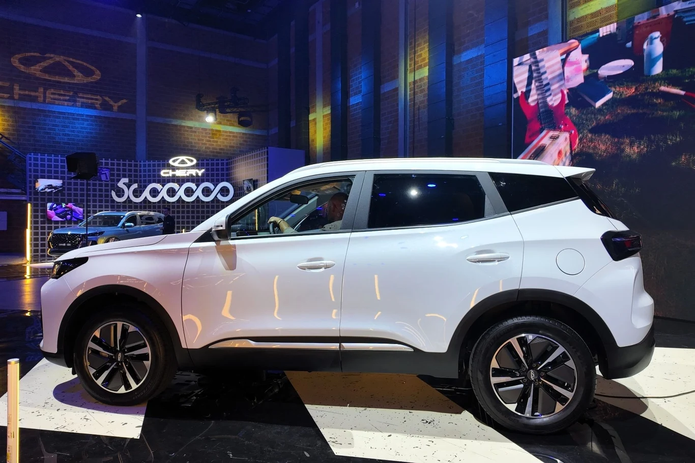 Chery Tiggo 4 HEV ma 4 320 mm, co oznacza, że jest konkurentem m.in. Volkswagena T-Roca. Biały samochód typu SUV ustawiony na czarnym błyszczącym podłożu w nowoczesnej hali wystawienniczej, w tle podświetlone logo marki Chery i ekran LED prezentujący inne elementy ekspozycji.