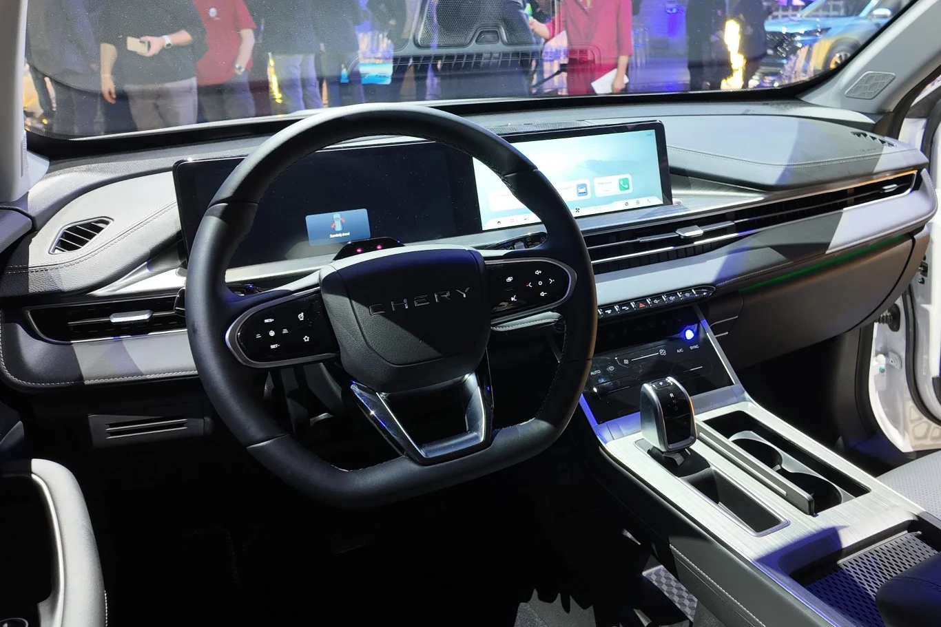 Centralnym punktem wnętrza Chery Tiggo 4 HEV są dwa 12,3-calowe wyświetlacze: wskaźników i multimediów. Kokpit nowoczesnego samochodu marki Chery z cyfrowymi wyświetlaczami, kierownicą wielofunkcyjną oraz zaawansowaną konsolą centralną z automatyczną skrzynią biegów; w tle rozmyte sylwetki ludzi.
