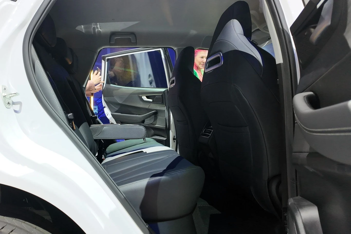 Z tyłu, jak na rozmiary Tiggo 4 HEV, jest sporo miejsca. Wnętrze samochodu widoczne od strony tylnych drzwi, siedzenia tapicerowane tkaniną i skórą, uchylone przednie oparcie umożliwia łatwiejsze wejście na tylną kanapę, na zewnątrz widać osoby przez otwarte drzwi.