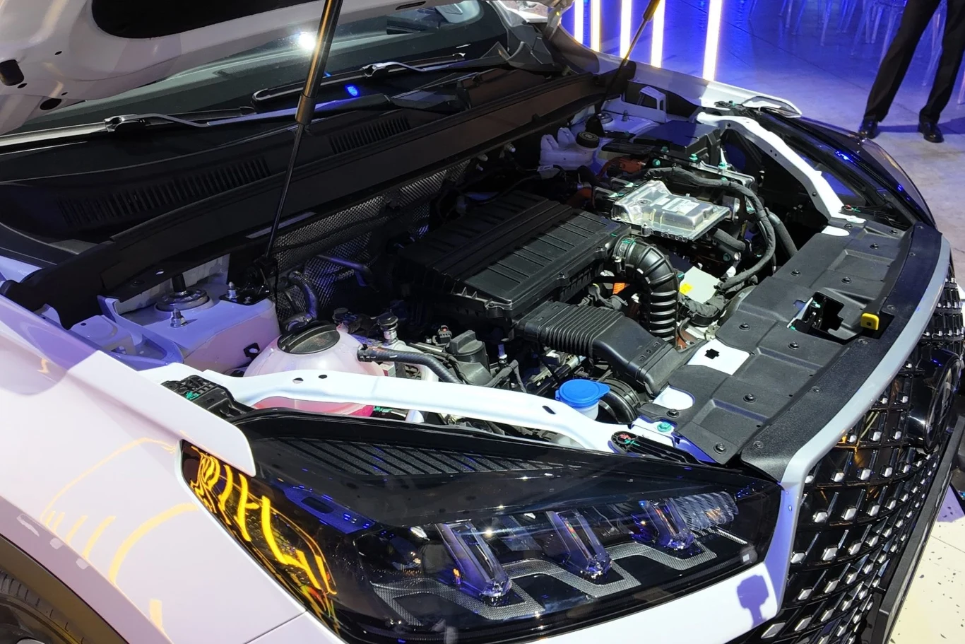 Hybrydowy układ Chery Tiggo 4 HEV oferuje systemowo 163 KM i 260 Nm. Otwarta maska samochodu odsłaniająca silnik i elementy układu mechanicznego, widoczne reflektory LED oraz detaliczna kratka wlotu powietrza, otoczenie nowoczesnego salonu samochodowego.