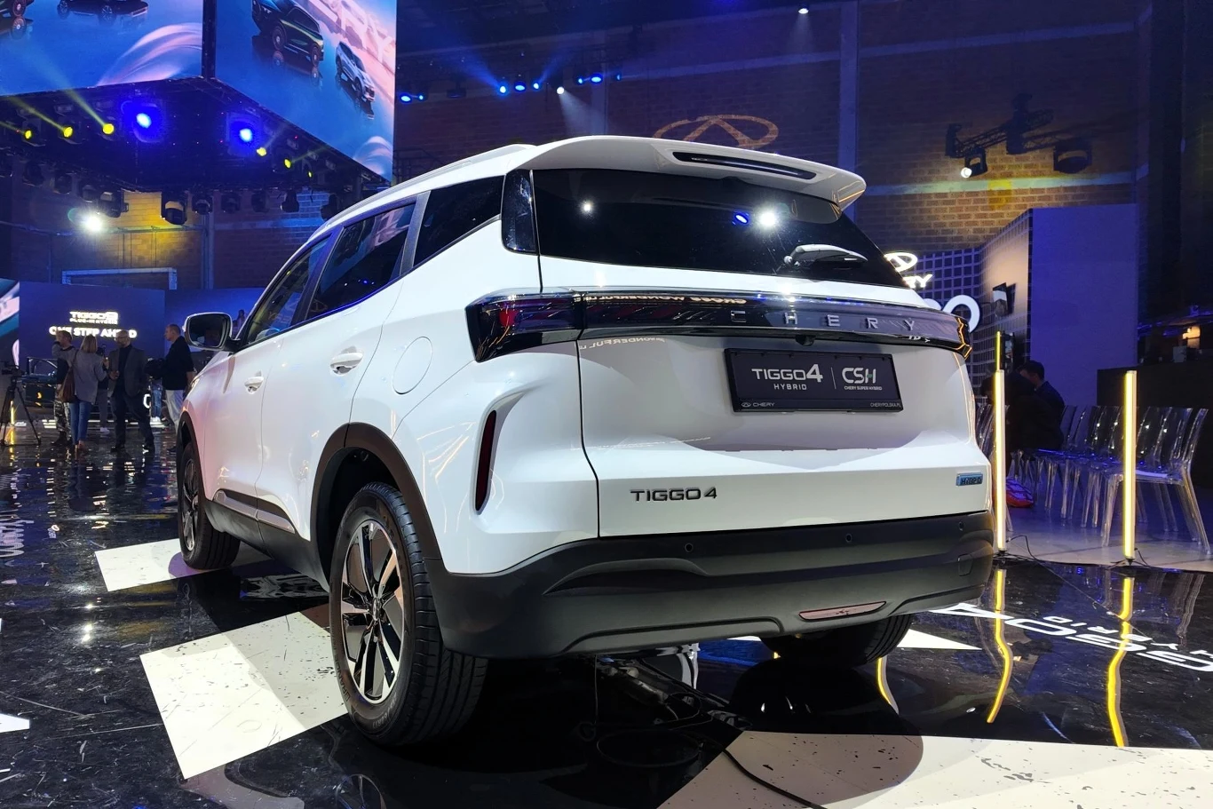 Chery Tiggo 4 HEV kosztuje minimum 95 900 zł. Biały samochód typu SUV marki Chery Tiggo 4 wystawiony na oświetlonym stoisku podczas targów motoryzacyjnych, widok od tyłu, wokół scena targowa z reflektorami i widoczne elementy wystroju.