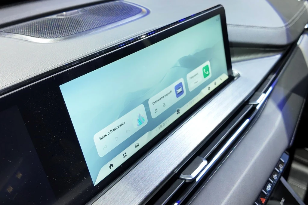 Duży ekran dotykowy systemu multimedialnego zamontowany w desce rozdzielczej samochodu, na którym wyświetlane są cyfrowe ikony funkcji oraz komunikaty z systemów pojazdu.