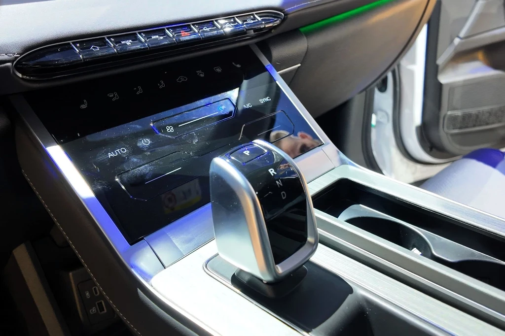 Nowoczesna konsola środkowa samochodu z elektroniczną dźwignią zmiany biegów, cyfrowym panelem sterowania klimatyzacją i dotykowymi przyciskami, w tle widoczne fragmenty wnętrza auta.