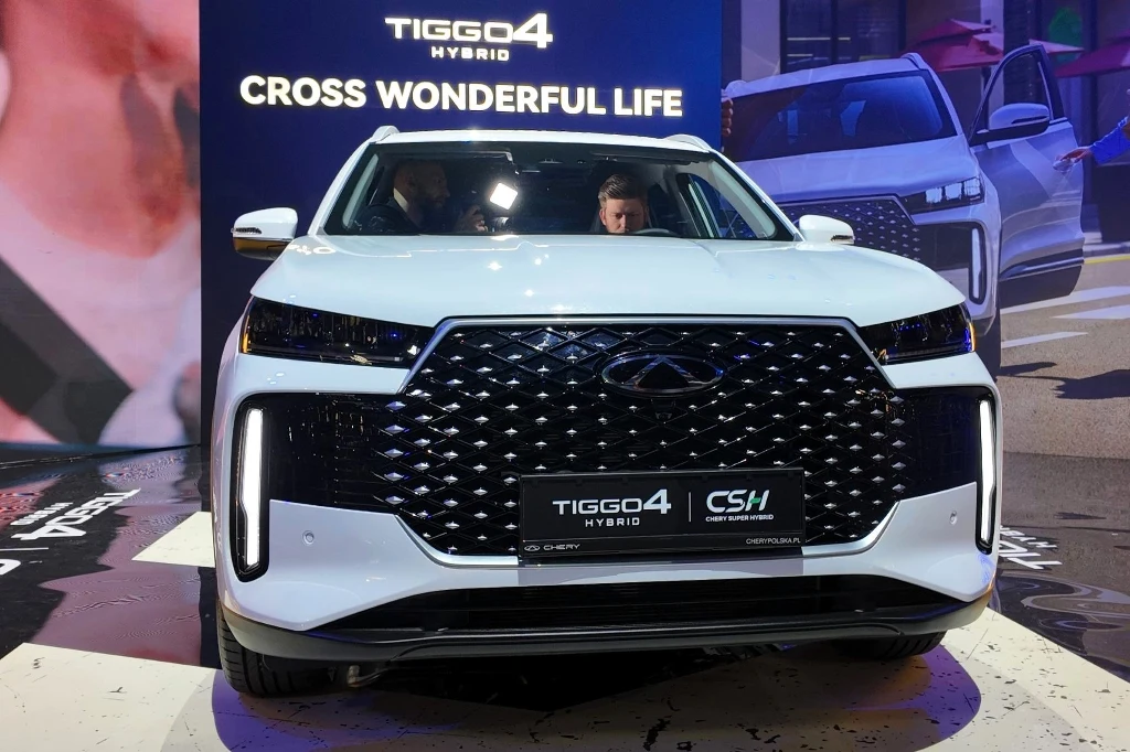 Nowoczesny biały samochód terenowy marki Chery Tiggo 4 Hybrid prezentowany na wystawie motoryzacyjnej, z dużym czarnym grillem, światłami LED i widocznym logo, w tle reklama z nazwą modelu oraz sloganem.