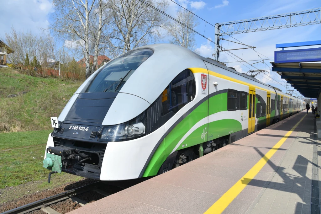 Koleje Mazowieckie dojadą do lotniska w Modlinie za cztery lata. Nowoczesny pociąg pasażerski w zielono-białych barwach stoi na peronie stacji kolejowej, w tle widoczne trakcje elektryczne, wiosenne drzewa i fragmenty infrastruktury kolejowej.