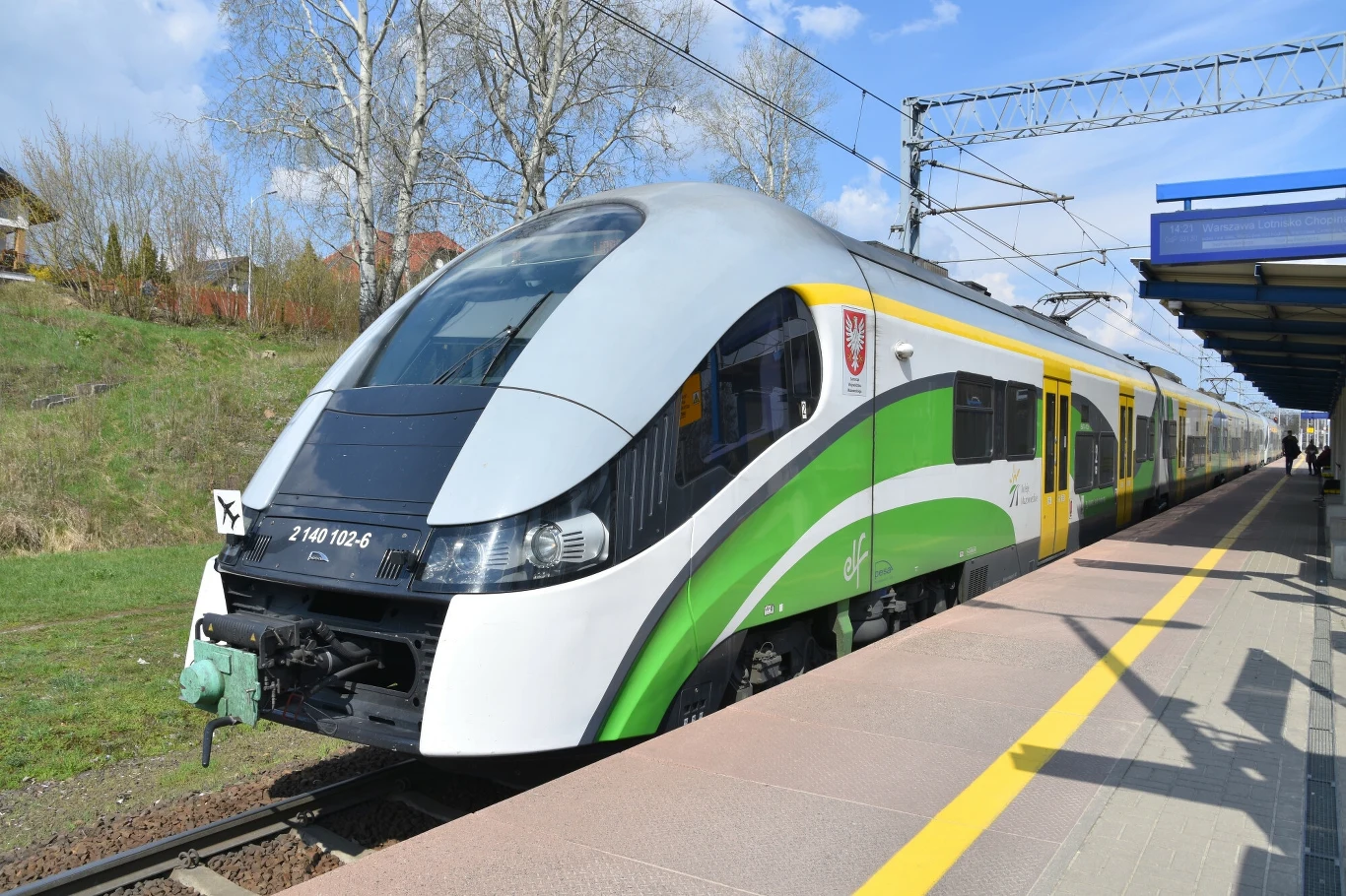 Nowoczesny pociąg pasażerski w zielono-białych barwach stoi na peronie stacji kolejowej, w tle widoczne trakcje elektryczne, wiosenne drzewa i fragmenty infrastruktury kolejowej.