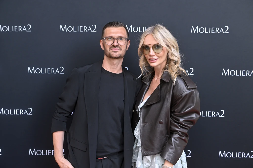 Dwoje eleganckich dorosłych pozuje razem na tle czarnej ścianki z nazwą Moliera2, mężczyzna ubrany w ciemną marynarkę i koszulkę, kobieta w skórzanej kurtce, jasnych okularach przeciwsłonecznych oraz jasnej sukience.