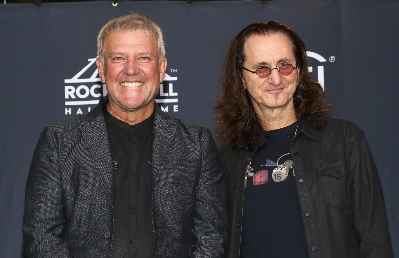Alex Lifeson i Geddy Lee (Rush) w Rock & Roll Hall Of Fame w 2017 r. Dwóch uśmiechniętych mężczyzn stoi na tle ścianki z logotypem Rock and Roll Hall of Fame, jeden z nich ma krótko ostrzyżone siwe włosy i ciemny garnitur, drugi długie ciemne włosy, okulary i ciemną koszulę.
