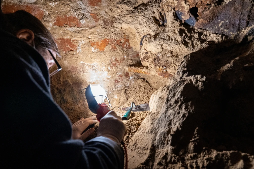 Otwarcie zamurowanej od lat krypty pod kompleksem klasztornym w Kazimierzu Biskupim przyniosło zaskakujące odkrycia. Osoba z latarką i narzędziem w ręku prowadzi prace badawcze lub wykopaliskowe w podziemnym tunelu z ceglaną ścianą i ziemistą powierzchnią.