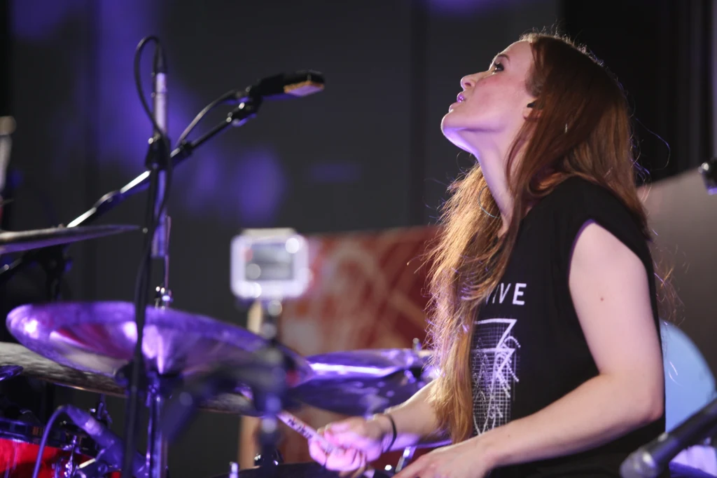 Kobieta z długimi włosami gra na perkusji, skupiona na muzyce, w tle widoczne elementy sceny i mikrofon.